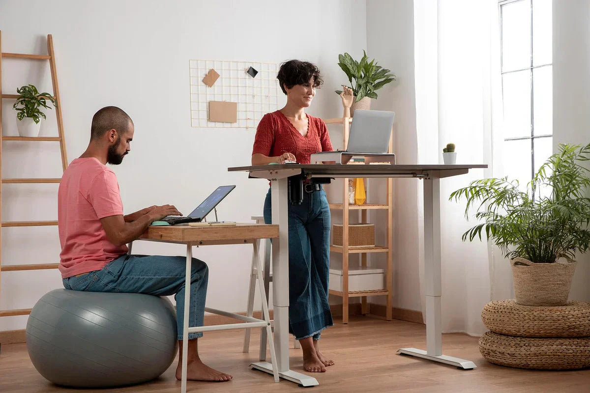 Man working on a laptop while sitting on an ergonomic Bloon chair and woman standing at a height-adjustable desk in a bright home office