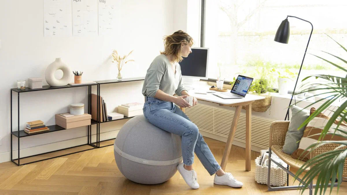Woman sitting on a gray ergonomic ball chair while working at a desk in a bright home office