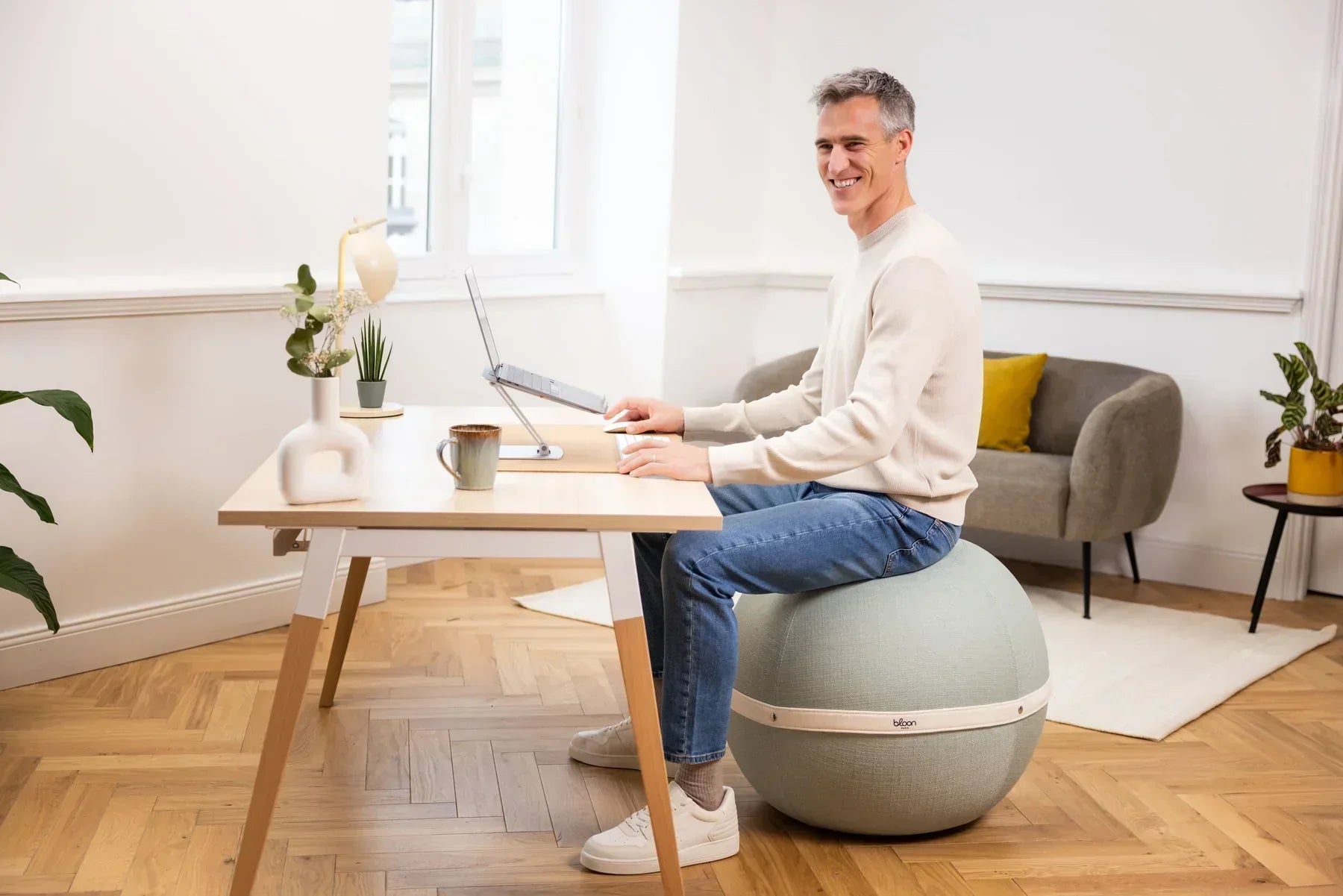 Man smiling while working on a laptop at a desk, seated on a light gray Bloon ergonomic ball chair in a modern office